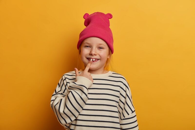 Horizontal shot of pretty small female child indicates at her new tooth, cares about teeth, wears stylish clothes, has funny expression, chuckles indoor, isolated over bright yellow background