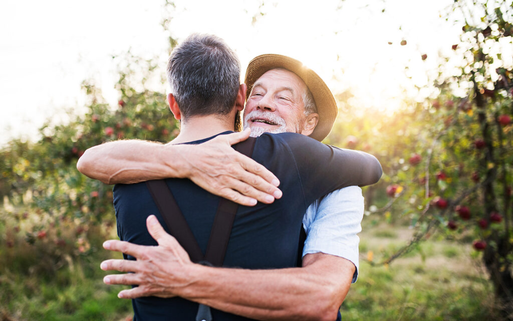 A senior man and adult son standing in apple orchard in autumn, hugging.