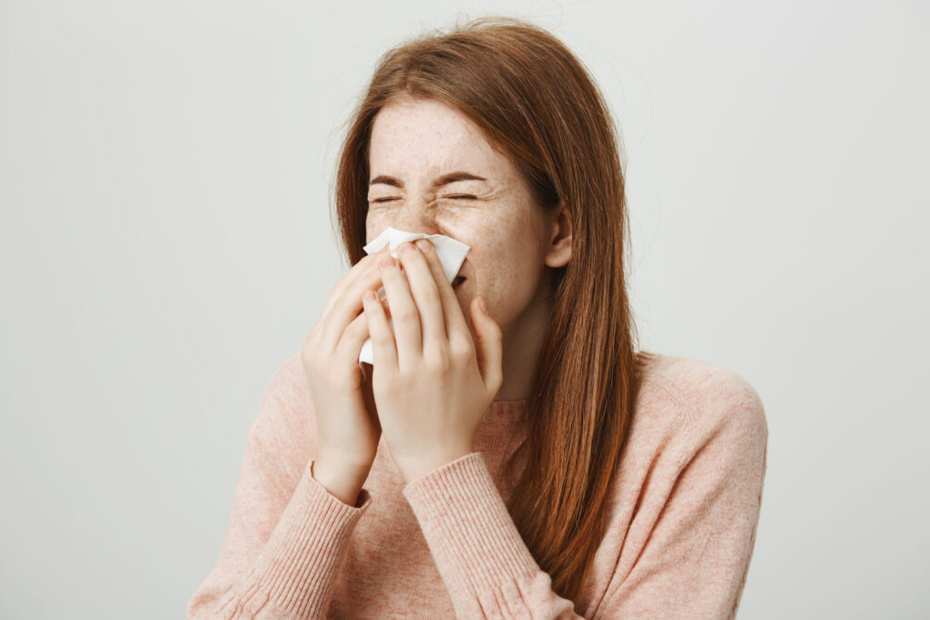 Close-up portrait of cute young ginger girl sneezing in napkin while standing against gray background. Woman has flu so she can not go to work today. Student crying while watching melodrama