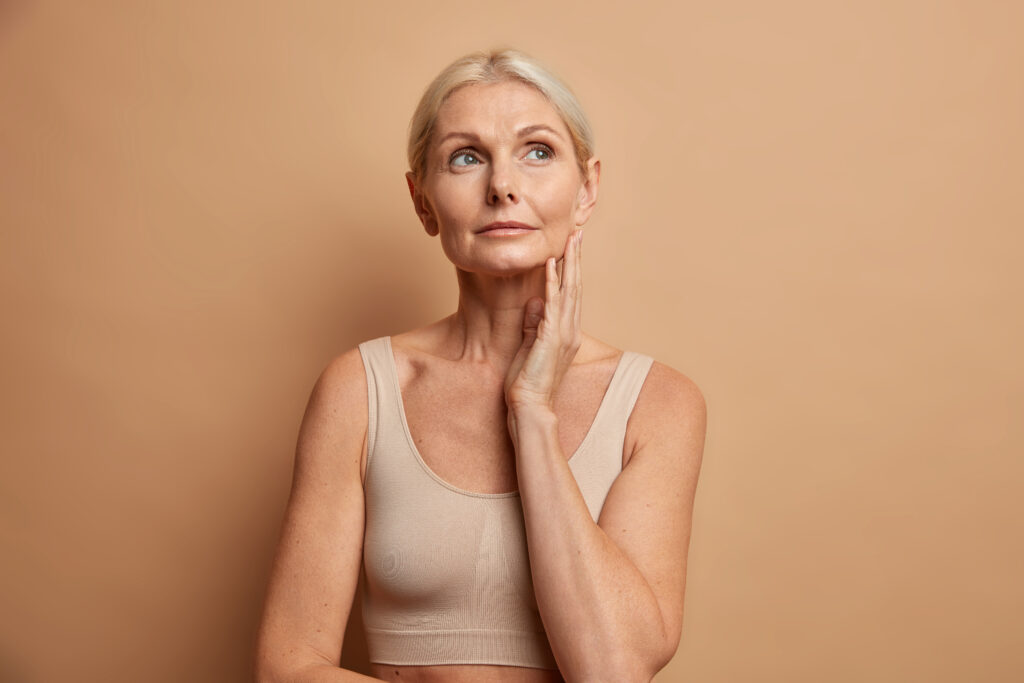 Serious tender wrinkled woman touches skin after applying anti age cream concentrated above with thoughtful expression wears cropped top isolated over brown background. Wellness hydration wellbeing Serious tender wrinkled woman touches skin after applying anti age cream concentrated above with thoughtful expression wears cropped top isolated over brown background. Wellness hydration wellbeing