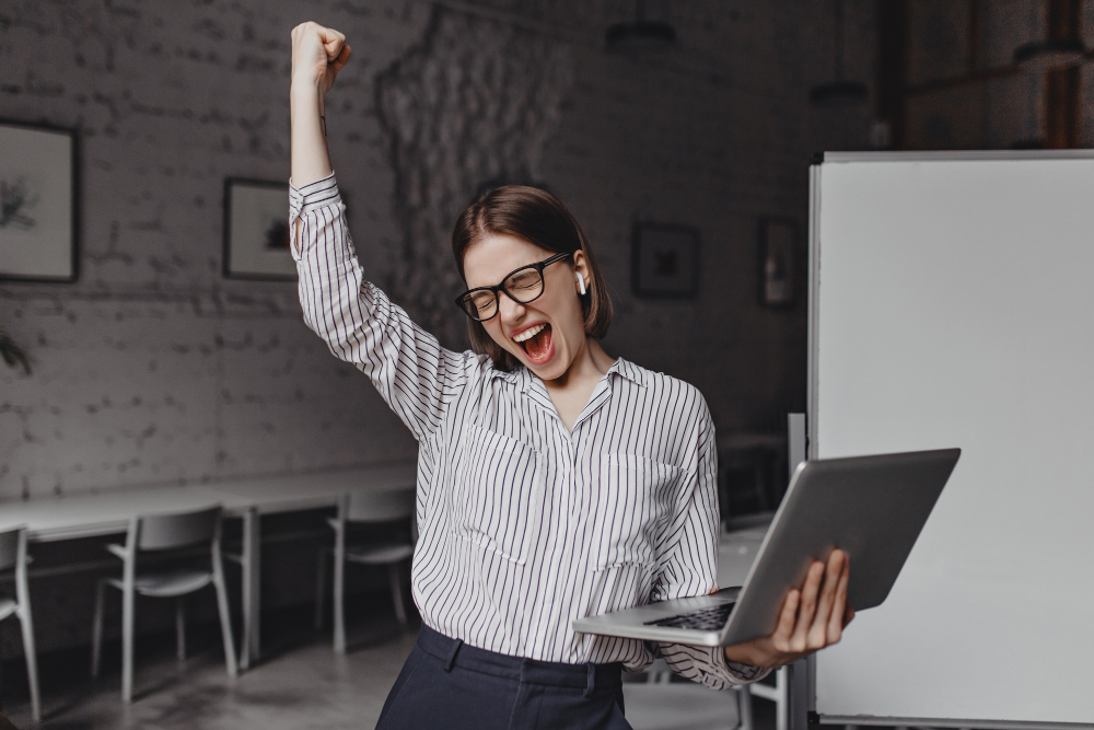 business-woman-with-laptop-in-hand-is-happy-with-success-portrait-of-woman-in-glasses-and-striped-blouse-enthusiastically-screaming-and-making-winning-gesture