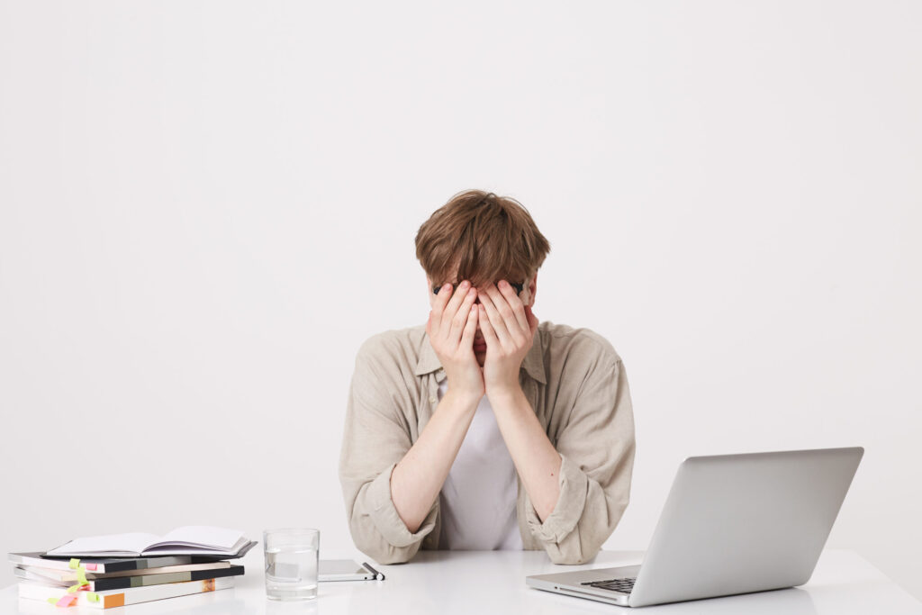 Closeup of tired desperate young man student wears beige shirt coveres face by hands sitting at the table with laptop computer and notebooks isolated over white background Closeup of tired desperate young man student wears beige shirt coveres face by hands sitting at the table with laptop computer and notebooks isolated over white background