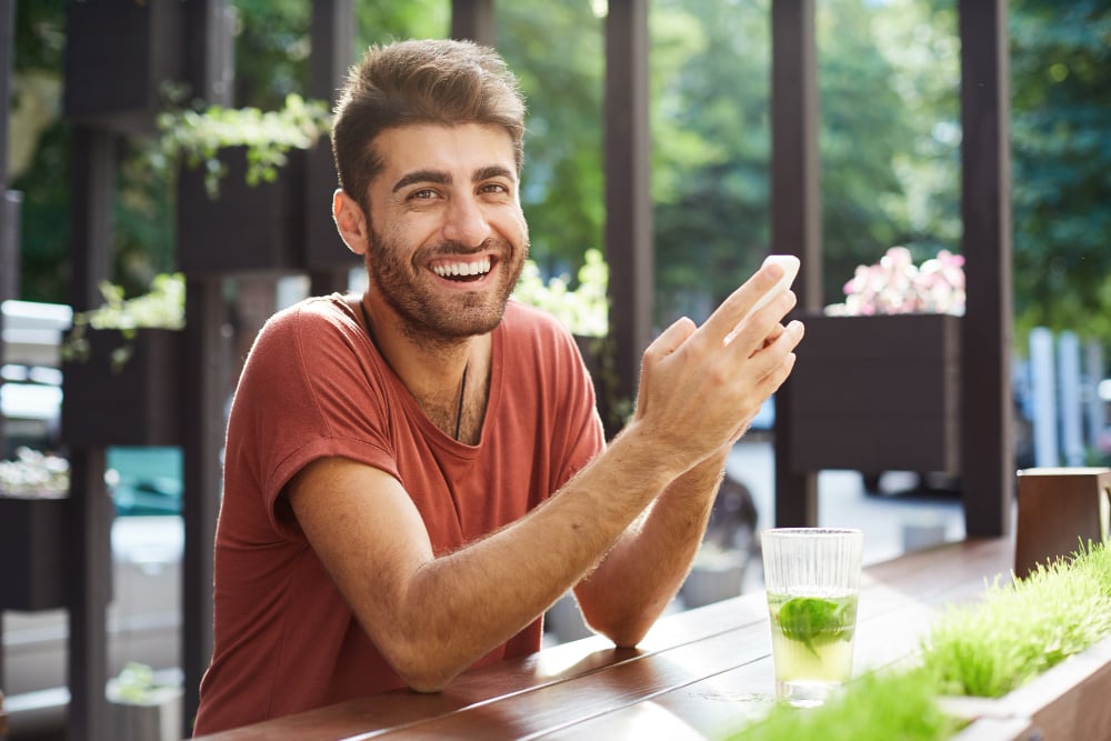 handsome-happy-guy-sitting-in-cafe-drinking-lemonade-and-using-mobile-phone-laughing-over-text-message