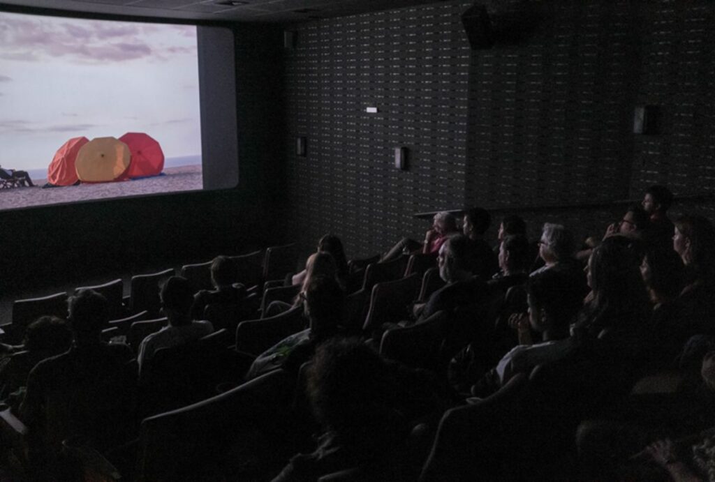 Sala de cinema escura com pessoas assistindo a um filme.