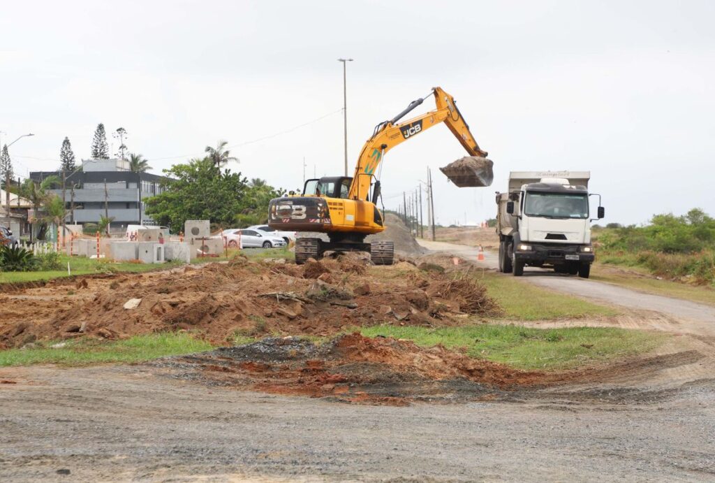 Escavadeira em terreno com caminhão ao fundo.