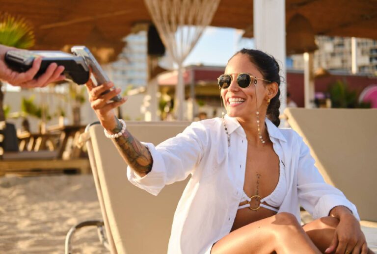 Mulher usando celular para fazer pagamento por aproximação sentada na praia.