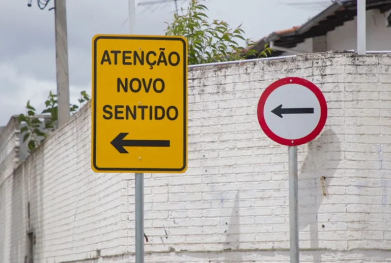 Placa de "atenção novo sentido" em frente a muro branco.