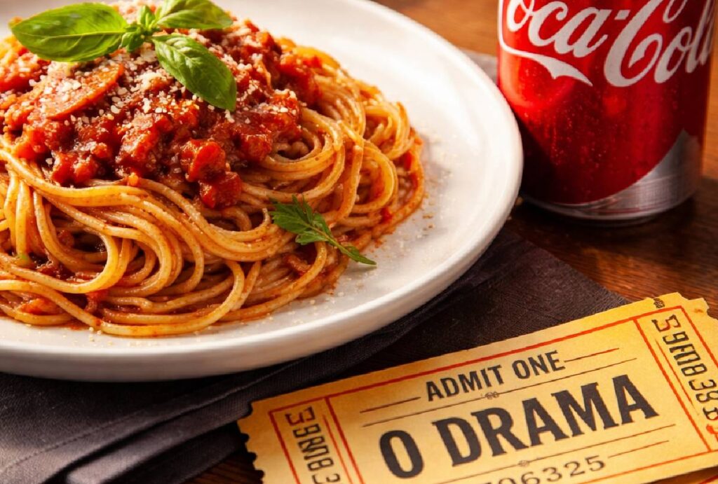 Prato de macarrão, ao lado de coca cola, com ingresso para "O Drama"
