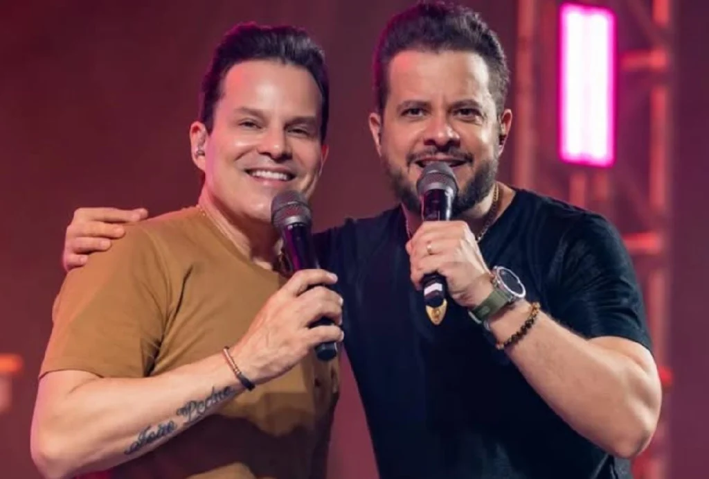 Dupla João Neto e Frederico com microfone em mãos sorrindo pra câmera