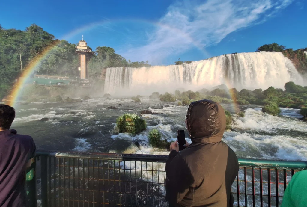 Turista tirando foto das Cataratas do Iguaçu em um dia de sol, enquanto um arco-íris se forma