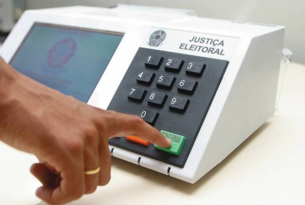 Mão clicando em confirmar em uma urna ELEITORAL.
